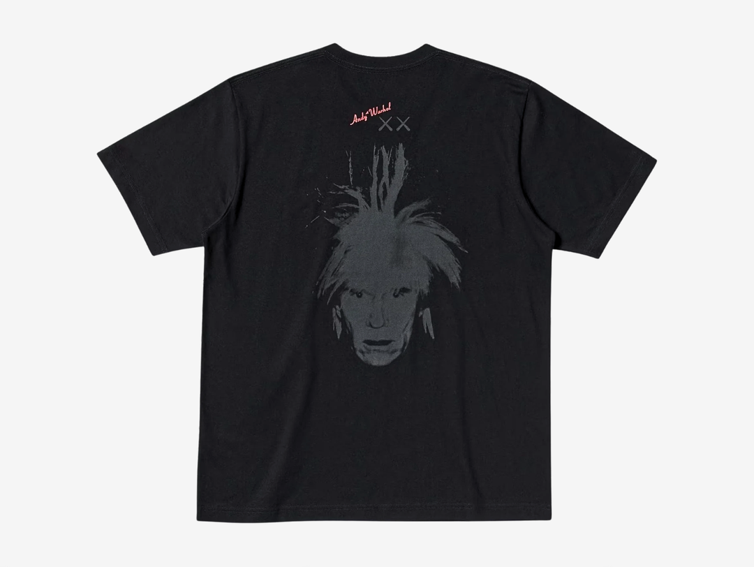 Black crewneck T-shirt, back view, featuring a red 'Andy Warhol' signature logo with grey KAWS 'XX' eyes printed below it, above a large tonal gray print of Andy Warhol's spiky self-portrait face, centered on the upper back.