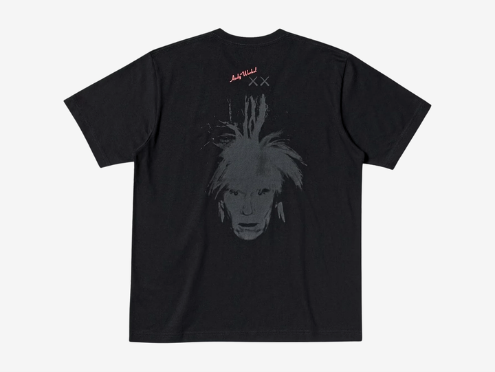 Black crewneck T-shirt, back view, featuring a red 'Andy Warhol' signature logo with grey KAWS 'XX' eyes printed below it, above a large tonal gray print of Andy Warhol's spiky self-portrait face, centered on the upper back.